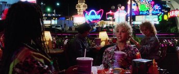Movie still from “Showgirls” (1995), directed by Paul Verhoeven – A woman sitting at a table with a drink in front of a neon sign; Medium shot, Over the shoulder angle