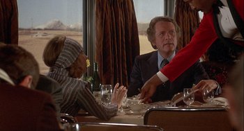 Movie still from “Silver Streak” (1976), directed by Arthur Hiller – A man sitting at a dinner table with two other people; Medium shot, Over the shoulder angle