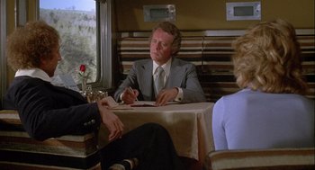 Movie still from “Silver Streak” (1976), directed by Arthur Hiller – A man sitting at a dinner table with two other people; Medium shot, Over the shoulder angle