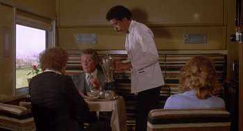 Movie still from “Silver Streak” (1976), directed by Arthur Hiller – A man standing at a table in front of two other people; Medium shot, High angle