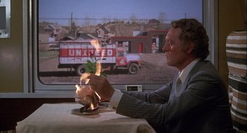 Movie still from “Silver Streak” (1976), directed by Arthur Hiller – A man sitting at a dinner table with a fire in his hand; Medium shot, High angle