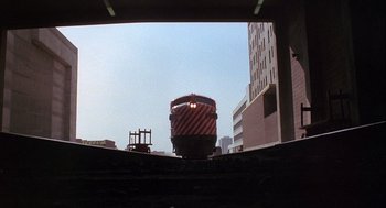 Movie still from “Silver Streak” (1976), directed by Arthur Hiller – A red and white train traveling down train tracks near buildings; Extreme Wide shot, Low angle