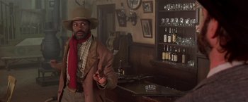 Movie still from “Silverado” (1985), directed by Lawrence Kasdan – A man standing in front of a bar with a bottle; Medium shot, Low angle