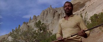 Movie still from “Silverado” (1985), directed by Lawrence Kasdan – A man standing in front of a rock formation holding a wooden stick; Medium shot, Low angle