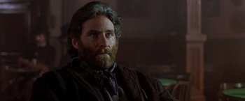 Movie still from “Silverado” (1985), directed by Lawrence Kasdan – A man with long hair and a beard is sitting in front of a window; Close Up shot, Low angle