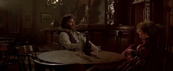 Movie still from “Silverado” (1985), directed by Lawrence Kasdan – A man sitting at a table with a bottle of alcohol; Medium shot, Over the shoulder angle