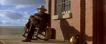 Movie still from “Silverado” (1985), directed by Lawrence Kasdan – A man sitting in a rocking chair on the porch of a brick building; Wide shot, Low angle