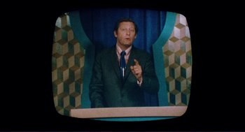 Movie still from “Sisters” (1972), directed by Brian De Palma – A man in a suit and tie standing in front of a microphone; Medium shot, High angle
