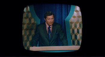 Movie still from “Sisters” (1972), directed by Brian De Palma – A man wearing a suit and tie sitting in front of a microphone; Medium shot, High angle