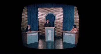 Movie still from “Sisters” (1972), directed by Brian De Palma – Three people sitting at a podium in front of a microphone; Extreme Wide shot, High angle