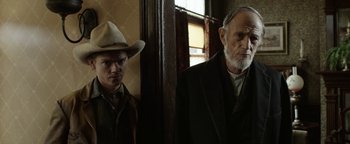 Movie still from “Godless” (2017), directed by Scott Frank – A man and a man with a hat standing next to each other in a room; Medium shot, Low angle