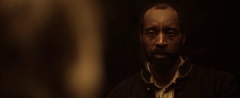 Movie still from “Godless” (2017), directed by Scott Frank – A man with a beard is standing in a dark room; Close Up shot, Over the shoulder angle