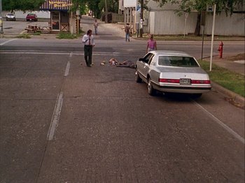 Movie still from “Slacker” (1990), directed by Richard Linklater – A man walking down the street next to a silver car; Wide shot, High angle