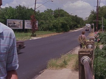 Movie still from “Slacker” (1990), directed by Richard Linklater – A man standing on the side of a road next to trees; Wide shot, Over the shoulder angle