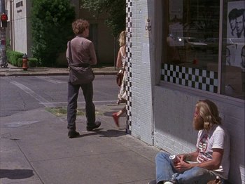 Movie still from “Slacker” (1990), directed by Richard Linklater – A man sitting on the side of the street next to a woman; Wide shot, Over the shoulder angle
