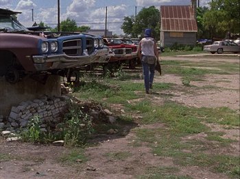 Movie still from “Slacker” (1990), directed by Richard Linklater – A man walking down a dirt road near a pile of old cars; Wide shot, Low angle