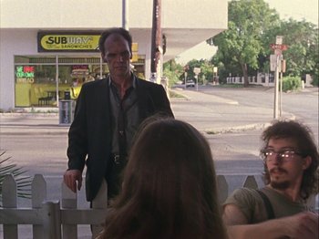 Movie still from “Slacker” (1990), directed by Richard Linklater – A man standing in front of a group of people; Medium shot, Over the shoulder angle