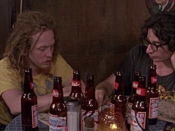 Movie still from “Slacker” (1990), directed by Richard Linklater – Two people sitting at a table with many beer bottles; Medium shot, High angle