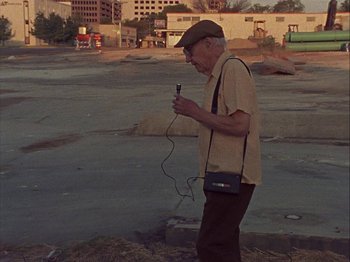 Movie still from “Slacker” (1990), directed by Richard Linklater – An older man is walking in a parking lot while using a cell phone; Wide shot, High angle