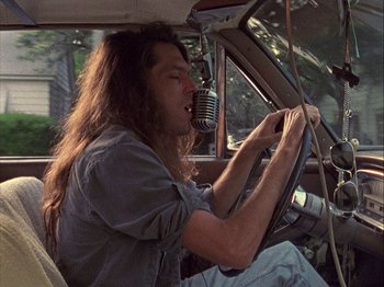 Movie still from “Slacker” (1990), directed by Richard Linklater – A man sitting in a car with a microphone in his hand; Medium shot, Low angle