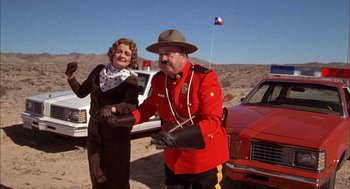 Movie still from “Smokey and the Bandit II” (1980), directed by Hal Needham – A man in a red jacket and a woman in a brown dress stand in front of a red car; Medium shot, Low angle