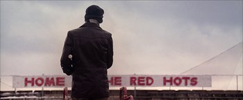 Movie still from “Snow Angels” (2007), directed by David Gordon Green – A man standing in front of a sign that reads " the red hot "; Wide shot, Low angle