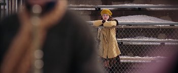 Movie still from “Snow Angels” (2007), directed by David Gordon Green – A woman in a yellow coat and hat leaning on a fence; Medium shot, Over the shoulder angle