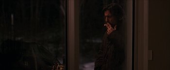 Movie still from “Snow Angels” (2007), directed by David Gordon Green – A man smoking a cigarette in front of a window at night; Medium shot, Over the shoulder angle