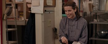 Movie still from “Snow Angels” (2007), directed by David Gordon Green – A young woman smiles while standing next to a wall; Medium shot, Over the shoulder angle