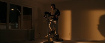Movie still from “Snow Angels” (2007), directed by David Gordon Green – A man standing on an exercise bike in a room; Wide shot, Low angle
