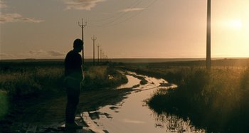 Movie still from “The Snowtown Murders” (2011), directed by Justin Kurzel – A person standing on a dirt road near a body of water; Extreme Wide shot, Low angle