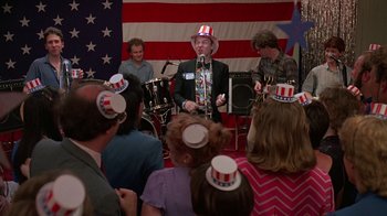 Movie still from “Something Wild” (1986), directed by Jonathan Demme – A group of people are gathered around a man in a patriotic hat; Medium shot, High angle