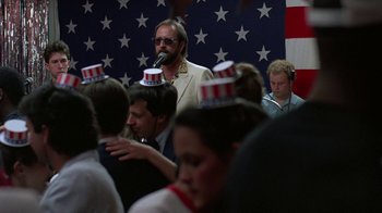 Movie still from “Something Wild” (1986), directed by Jonathan Demme – A group of people sitting in front of an american flag wall; Medium shot, Low angle