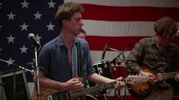 Movie still from “Something Wild” (1986), directed by Jonathan Demme – A man playing a guitar in front of an american flag; Medium shot, Low angle
