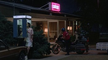 Movie still from “Something Wild” (1986), directed by Jonathan Demme – A police officer on a motorcycle in front of a house; Wide shot, Over the shoulder angle