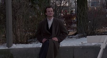 Movie still from “Groundhog Day” (1993), directed by Harold Ramis – A man wearing a long coat sitting on a wall; Wide shot, Low angle