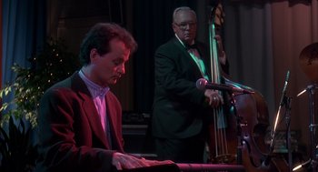 Movie still from “Groundhog Day” (1993), directed by Harold Ramis – A man in a suit playing a piano while a man in a suit plays a double bass; Medium shot, Over the shoulder angle