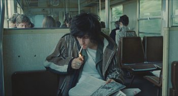 Movie still from “Something in the Air” (2012), directed by Olivier Assayas – A man sitting on a bus smoking a cigarette and reading a book; Medium shot, High angle