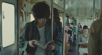 Movie still from “Something in the Air” (2012), directed by Olivier Assayas – A man reading a book while riding on a bus; Medium shot, Over the shoulder angle