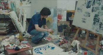 Movie still from “Something in the Air” (2012), directed by Olivier Assayas – A man sitting on the floor in a room with many paint brushes; Medium shot, High angle