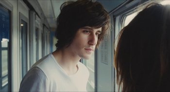 Movie still from “Something in the Air” (2012), directed by Olivier Assayas – A man with long hair and a white t - shirt is looking out a window; Close Up shot, Over the shoulder angle