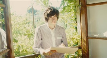 Movie still from “Something in the Air” (2012), directed by Olivier Assayas – A young man holding a laptop and writing on a piece of paper; Medium shot, Low angle