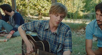Movie still from “Something in the Air” (2012), directed by Olivier Assayas – A man playing a guitar in the grass; Medium shot, Over the shoulder angle