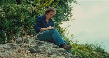 Movie still from “Something in the Air” (2012), directed by Olivier Assayas – A man sitting on top of a rock in the grass; Medium shot, Low angle