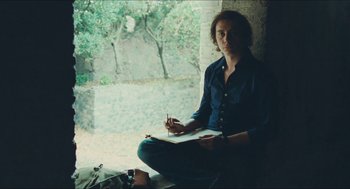 Movie still from “Something in the Air” (2012), directed by Olivier Assayas – A man sitting on the ground holding a book; Medium shot, Low angle