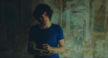 Movie still from “Something in the Air” (2012), directed by Olivier Assayas – A man in a blue t - shirt is holding a notebook and a pen; Medium shot, Overhead angle