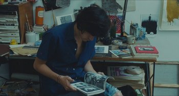 Movie still from “Something in the Air” (2012), directed by Olivier Assayas – A woman sitting in a room holding a book; Medium shot, Overhead angle