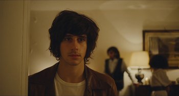 Movie still from “Something in the Air” (2012), directed by Olivier Assayas – A man with long hair and a white t - shirt is in a room; Close Up shot, Over the shoulder angle