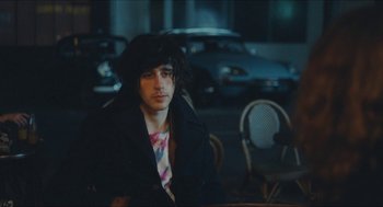 Movie still from “Something in the Air” (2012), directed by Olivier Assayas – A man sitting at a table in front of a row of cars; Medium shot, Over the shoulder angle