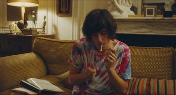 Movie still from “Something in the Air” (2012), directed by Olivier Assayas – A person sitting on a couch smoking a cigarette; Medium shot, Over the shoulder angle